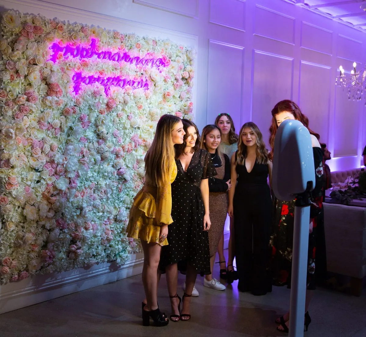 Friends posing in front of a flower wall with a pink neon sign